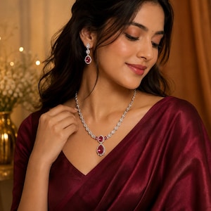 May include: A silver-toned necklace and matching drop earrings set featuring pear-shaped deep pink stones surrounded by small clear stones. The necklace has a central pear-shaped pendant and a chain composed of circular links with clear stones.