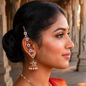 May include: A gold jhumka earring featuring alternating green and pink stones with small white pearls, connected to a matching ear chain that hooks into the hair. A small gold nose stud and a gold necklace with matching stones are also visible.