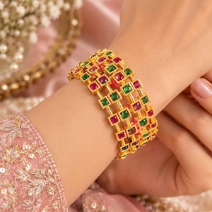 May include: A gold-toned bangle bracelet featuring a grid-like structure set with alternating square-cut red and green gemstones. The bracelet consists of three parallel rows of metal links, each containing small, vibrant, faceted stones.