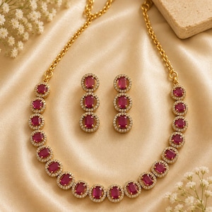May include: A gold-toned necklace and matching earring set featuring rectangular, deep pink stones surrounded by small, clear white crystals. The necklace consists of a chain of these stone settings, while each earring features three stacked stones.