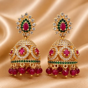May include: A pair of gold-tone jhumka earrings featuring a central teardrop-shaped red stone surrounded by green and clear crystals. The bell-shaped base is adorned with circular red stone accents, clear crystals, and dangling red beads.