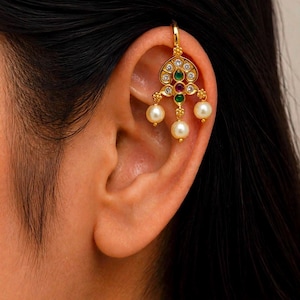 May include: A gold ear cuff featuring a decorative filigree design adorned with clear crystals, small green and red stones, and three dangling white pearls. The piece is shaped to hook onto the upper cartilage of the ear, showcasing a detailed metalwork.