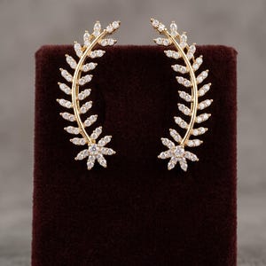 May include: A pair of gold-toned ear climbers shaped like curved laurel branches. Each earring features a central stem adorned with small, clear, sparkling stones arranged in a leaf pattern, culminating in a star-shaped cluster at the base.