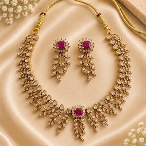 May include: A gold-toned necklace and matching earring set featuring a leaf-inspired design adorned with small clear stones and a central rectangular magenta stone. The necklace includes a thin gold cord with an adjustable sliding bead closure.