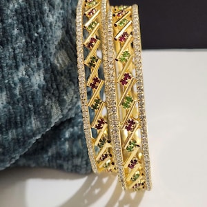 May include: Two gold bangles featuring a central lattice design set with alternating small green and red stones. Each bangle is bordered by two rows of tiny clear white stones along the edges, creating a structured and metallic appearance.