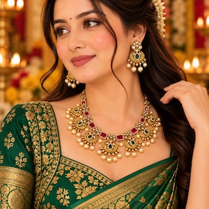 May include: A gold-toned necklace and matching drop earrings set featuring square red stones, teardrop-shaped green stones, and dangling white pearls. The jewelry is worn with a dark green blouse featuring a gold floral pattern and gold trim.