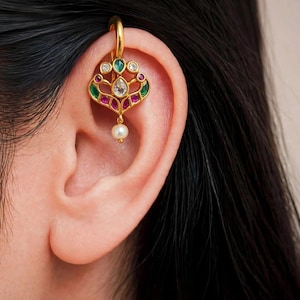 May include: A gold ear cuff featuring a decorative filigree design adorned with small green, red, and clear stones. The piece includes a central teardrop-shaped clear stone and a single dangling white pearl at the bottom, secured to the upper ear cartilage.