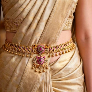 May include: A gold-toned waist belt featuring intricate metalwork, embedded with vibrant pink and green stones. The design includes repeating peacock-like motifs and a central floral-shaped pendant with dangling gold beads, worn over a beige fabric.