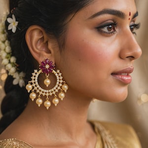 May include: A gold-toned chandelier earring featuring a central flower-shaped motif with deep pink petals, a circular frame encrusted with small clear stones, and multiple dangling cream-colored pearls arranged along the bottom edge.
