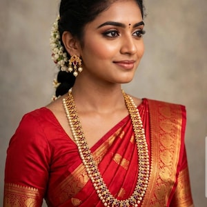 May include: A long gold necklace featuring intricate metalwork, small red stones, and dangling white pearls. Matching gold earrings with red stones and pearl drops are worn. The jewelry is paired with a red silk saree with gold woven borders.