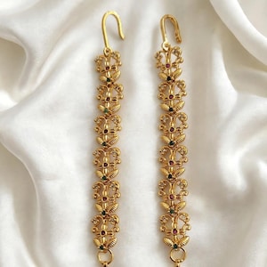 May include: A pair of long gold-toned metal ear chains featuring a repeating floral and leaf-inspired filigree pattern. Each chain is accented with small embedded red and green stones and terminates in a hook at the top and circular rings at the base.