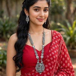 May include: A long silver-toned necklace featuring intricate metalwork and a large central pendant with a small green stone, paired with matching silver-toned drop earrings. The jewelry is worn with a deep red sleeveless top and a red patterned shawl.