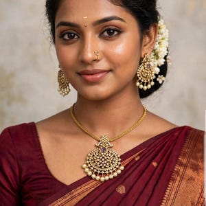 May include: A gold necklace featuring a large, circular pendant with intricate filigree, small white pearls, and a central purple stone, paired with matching gold chandelier earrings with pearls and a small gold nose ring with a purple stone.