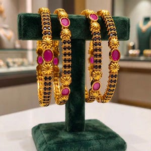 May include: A pair of gold bangles featuring alternating sections of small black stones and larger, oval-shaped pink stones. The gold metalwork includes textured, sunburst-like circular accents between the stones, creating a repeating pattern.