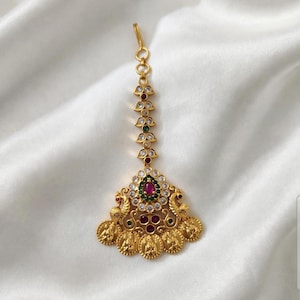 May include: A gold-toned maang tikka headpiece featuring a vertical chain of clear stones and small leaf-shaped accents. The pendant base displays two peacock figures, a central oval ruby-colored stone, green accents, and five hanging coin motifs.