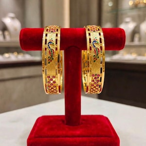 May include: A pair of gold bangles featuring a central peacock motif with blue, green, and red enamel detailing. The bangles have a textured gold surface and are accented with small, round red stones arranged in a repeating pattern along the edges.
