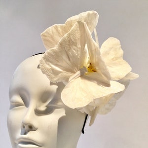 Puede incluir: Una diadema blanca con una flor de diseño delicado y superpuesto. La diadema está hecha de tela y tiene una banda elástica negra simple.