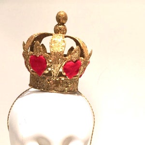 May include: A gold glitter crown headband featuring a central sphere topper and red fabric heart accents integrated into the crown structure. The crown is mounted on a thin, matching gold glitter headband, creating a cohesive headpiece design.