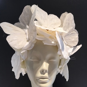 May include: A white headpiece made of paper butterflies. The butterflies are arranged in a circular pattern around the head. The headpiece is worn on a white mannequin head.