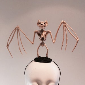 May include: A Halloween-themed headband featuring a skeletal bat figure. The bat is beige and stands atop a small black platform attached to a black headband. The figure has outstretched wings and a detailed skull.