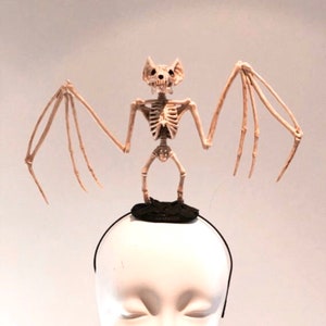May include: A Halloween-themed headband featuring a skeletal bat figure. The bat is beige and has outstretched wings, perched atop a black, circular base. The headband itself is black, and the display is on a mannequin head.
