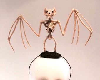May include: A Halloween-themed headband featuring a skeletal bat figure. The bat is beige and has outstretched wings, perched atop a black, circular base. The headband itself is black, and the display is on a mannequin head.