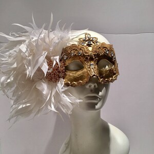 May include: A gold masquerade mask with white feathers and rhinestones. The mask is on a white mannequin head.