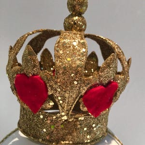 May include: A gold glitter-covered crown featuring a circular band and arched points topped with a small sphere. The crown is decorated with two prominent red velvet heart-shaped appliqu&eacute;s attached to the front, creating a textured appearance.