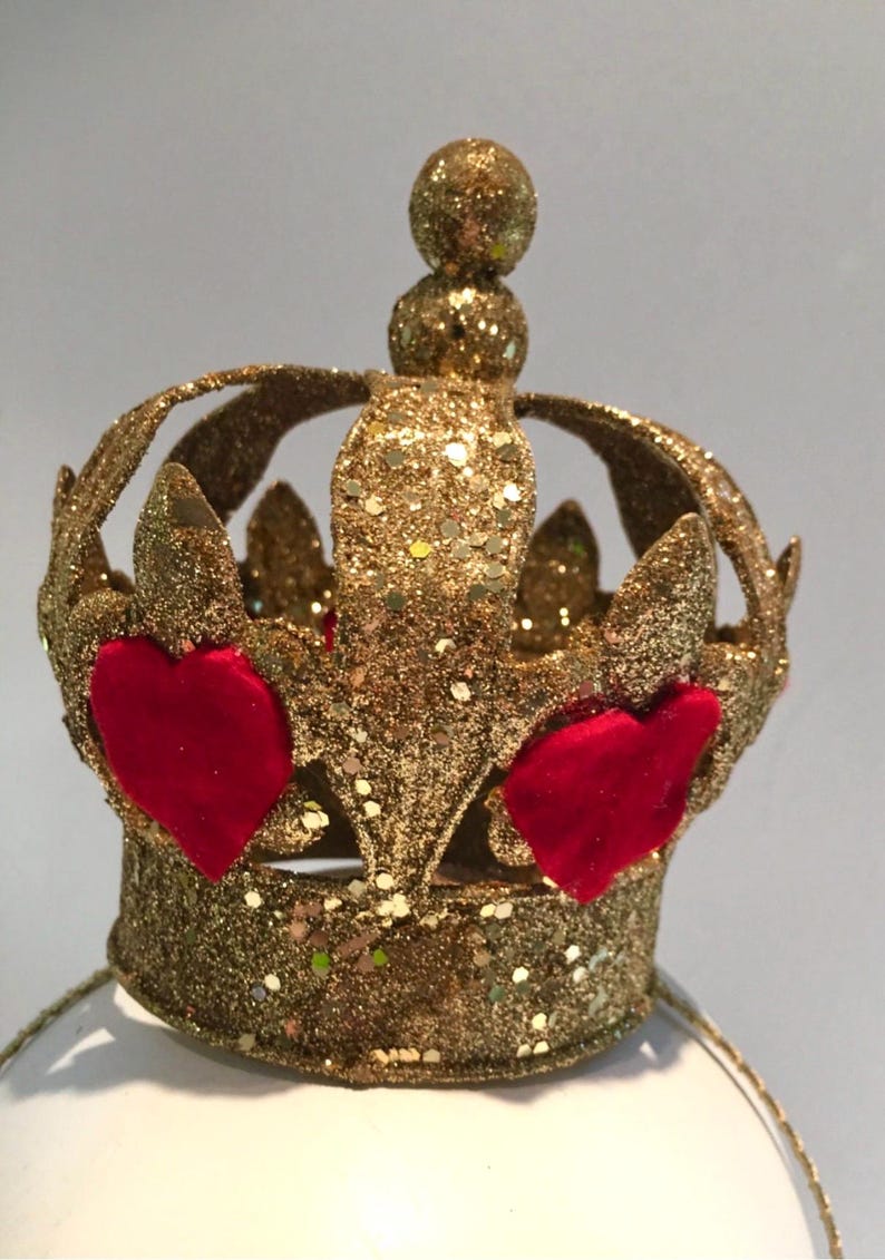 May include: A gold glitter-covered crown featuring a circular band and arched points topped with a small sphere. The crown is decorated with two prominent red velvet heart-shaped appliqu&eacute;s attached to the front, creating a textured appearance.