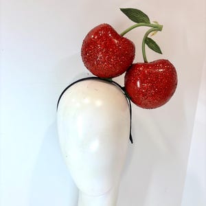 K&ouml;nnte beinhalten: Ein schwarzes Stirnband mit zwei gro&szlig;en, rot glitzernden Kirschen und gr&uuml;nen Bl&auml;ttern. Die Kirschen sind an einem gr&uuml;nen Stiel befestigt. Das Stirnband wird auf einem wei&szlig;en Schaufensterkopf pr&auml;sentiert.
