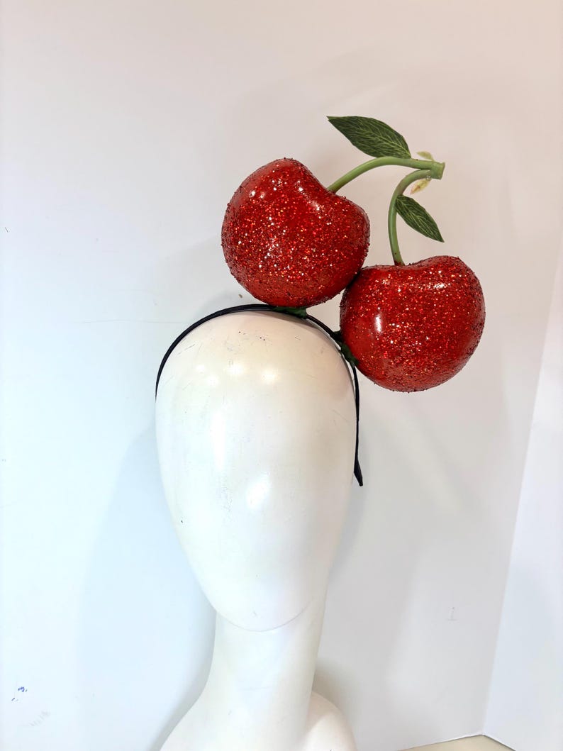 K&ouml;nnte beinhalten: Ein schwarzes Stirnband mit zwei gro&szlig;en, rot glitzernden Kirschen und gr&uuml;nen Bl&auml;ttern. Die Kirschen sind an einem gr&uuml;nen Stiel befestigt. Das Stirnband wird auf einem wei&szlig;en Schaufensterkopf pr&auml;sentiert.