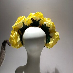 May include: A floral headband featuring a series of large, vibrant yellow artificial roses attached to a dark green base. The roses are arranged in a dense, circular pattern, showcasing layered petals and green foliage accents throughout the design.