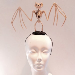 May include: A Halloween-themed headband featuring a skeletal bat figure. The bat is beige with outstretched wings, perched atop a black base attached to a thin black headband. The head of a mannequin is visible below the headband.