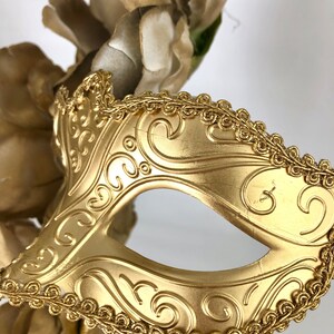 K&ouml;nnte beinhalten: Eine goldfarbene Maske f&uuml;r einen Maskenball mit einer Augen&ouml;ffnung und einem verzierten Rankenmuster. Die Maske ist mit einer goldfarbenen, seilartigen Bord&uuml;re versehen und vor einem Hintergrund aus goldfarbenen Kunstblumen platziert.