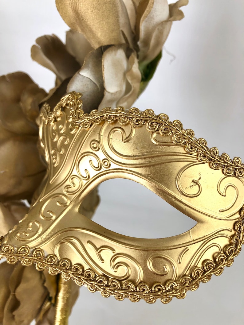 K&ouml;nnte beinhalten: Eine goldfarbene Maske f&uuml;r einen Maskenball mit einer Augen&ouml;ffnung und einem verzierten Rankenmuster. Die Maske ist mit einer goldfarbenen, seilartigen Bord&uuml;re versehen und vor einem Hintergrund aus goldfarbenen Kunstblumen platziert.