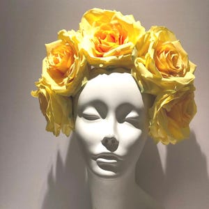 May include: A floral headpiece featuring a crown of large, vibrant yellow artificial roses arranged in a circular pattern. The flowers are attached to a base that rests atop a smooth, white, featureless mannequin head with closed eyes.
