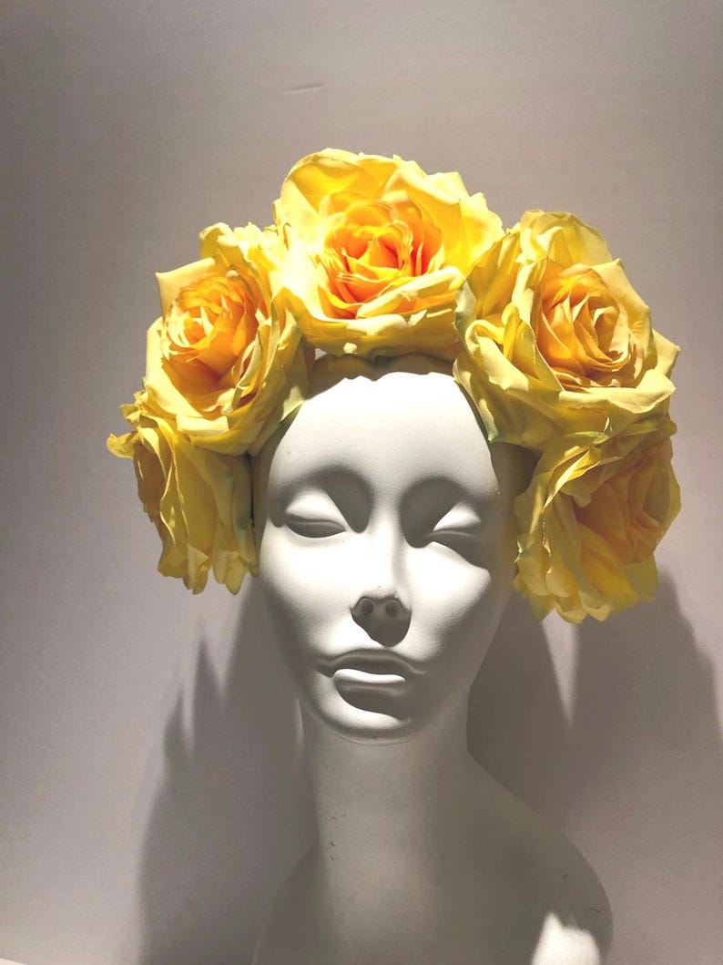 May include: A floral headpiece featuring a crown of large, vibrant yellow artificial roses arranged in a circular pattern. The flowers are attached to a base that rests atop a smooth, white, featureless mannequin head with closed eyes.