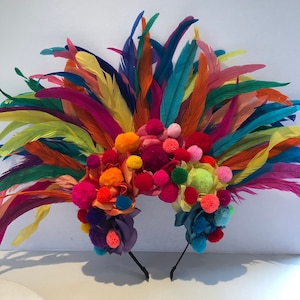 May include: A colorful headband with a large cluster of pom-poms in various shades of pink, orange, yellow, green, and purple. The headband is adorned with feathers in a rainbow of colors, including red, orange, yellow, green, blue, and purple.