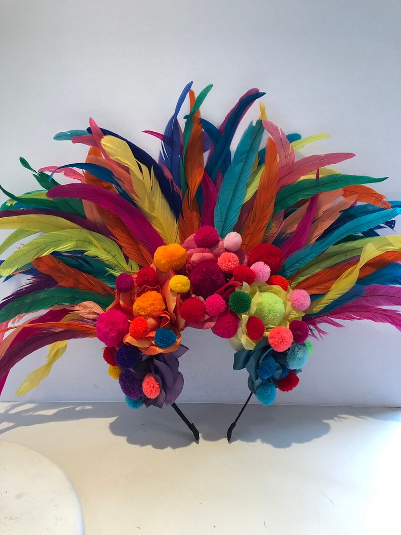 May include: A colorful headband with a large cluster of pom-poms in various shades of pink, orange, yellow, green, and purple. The headband is adorned with feathers in a rainbow of colors, including red, orange, yellow, green, blue, and purple.