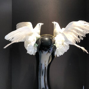 May include: A decorative black vase with a glossy finish, adorned with two white dove figurines. The doves have outstretched wings and are surrounded by white floral accents. The vase has a slender neck and a wide base.