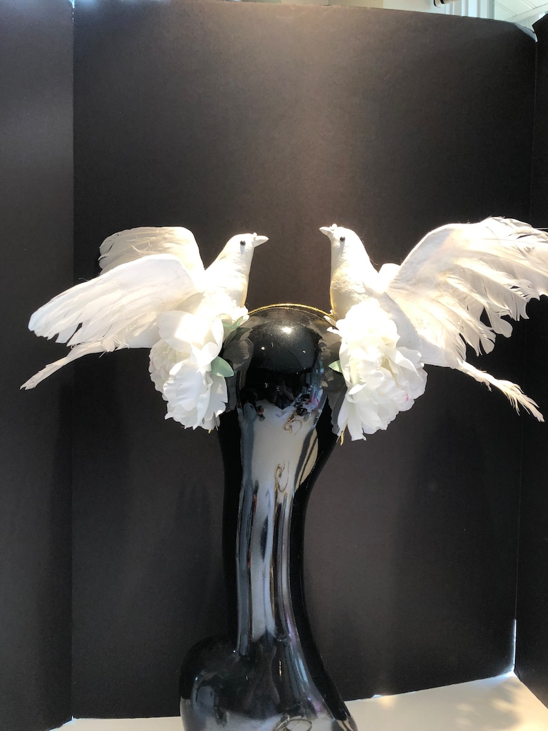 May include: A decorative black vase with a glossy finish, adorned with two white dove figurines. The doves have outstretched wings and are surrounded by white floral accents. The vase has a slender neck and a wide base.