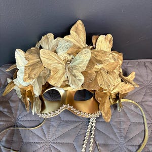 Puede incluir: Una m&aacute;scara dorada para un baile de m&aacute;scaras con detalles de mariposas. La m&aacute;scara presenta aberturas para los ojos, un ribete decorativo de pedrer&iacute;a y cintas doradas. La parte superior de la m&aacute;scara est&aacute; adornada con m&uacute;ltiples mariposas doradas.