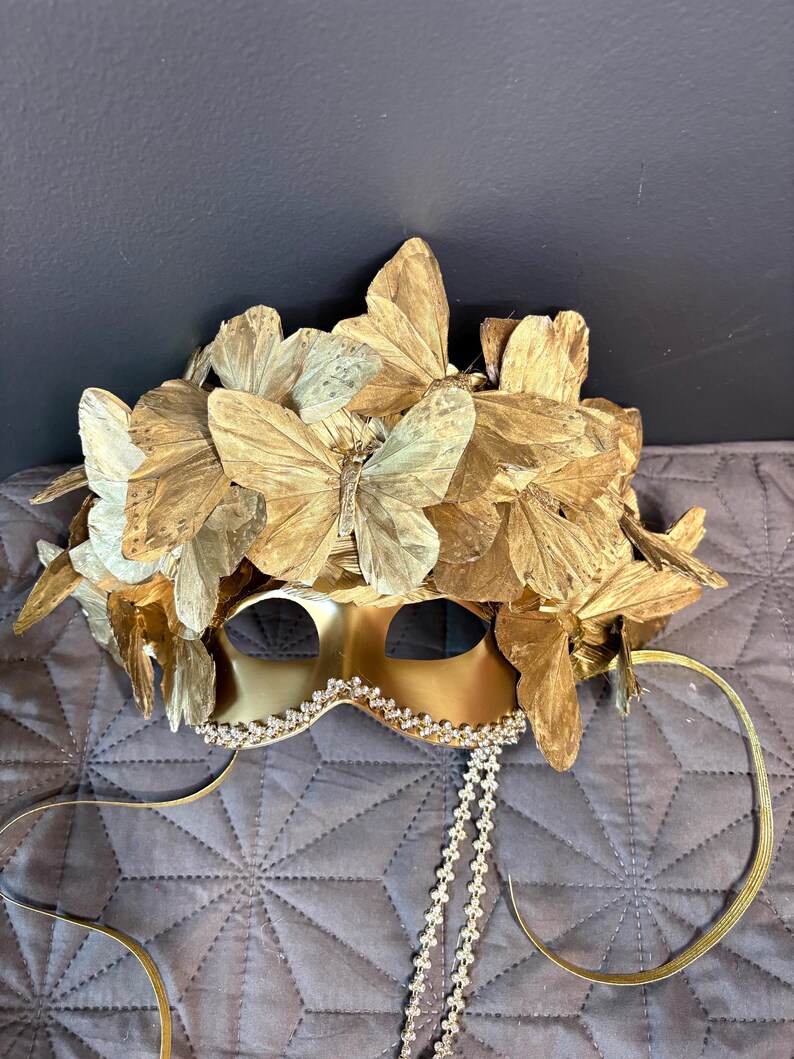 Puede incluir: Una m&aacute;scara dorada para un baile de m&aacute;scaras con detalles de mariposas. La m&aacute;scara presenta aberturas para los ojos, un ribete decorativo de pedrer&iacute;a y cintas doradas. La parte superior de la m&aacute;scara est&aacute; adornada con m&uacute;ltiples mariposas doradas.