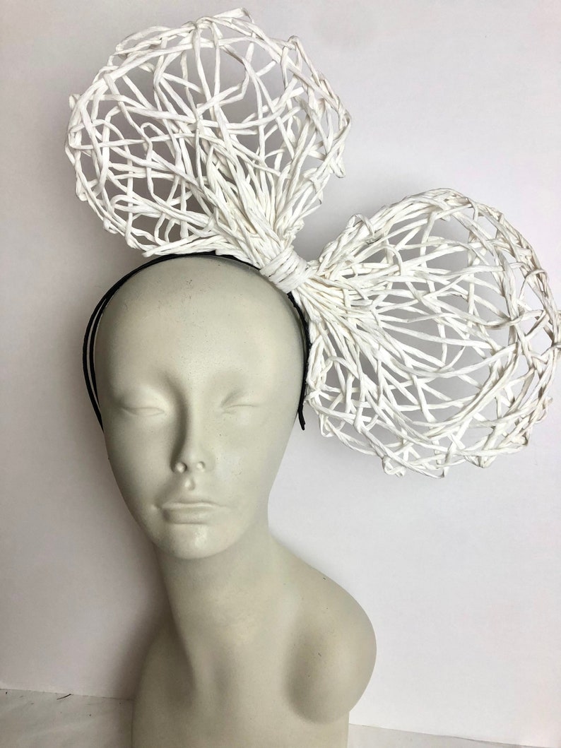 May include: A white woven headband with a large, white, woven bow. The bow is made of thin, white strips of material that are woven together to create a delicate, airy look.