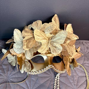 Puede incluir: Una m&aacute;scara dorada para carnaval con detalles de mariposas. La m&aacute;scara presenta un borde decorativo en el borde inferior y una cascada de peque&ntilde;as cuentas transparentes. Cintas doradas y alas de mariposa adornan la parte superior.
