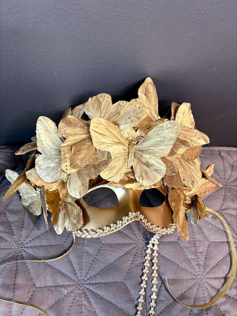 Puede incluir: Una m&aacute;scara dorada para carnaval con detalles de mariposas. La m&aacute;scara presenta un borde decorativo en el borde inferior y una cascada de peque&ntilde;as cuentas transparentes. Cintas doradas y alas de mariposa adornan la parte superior.