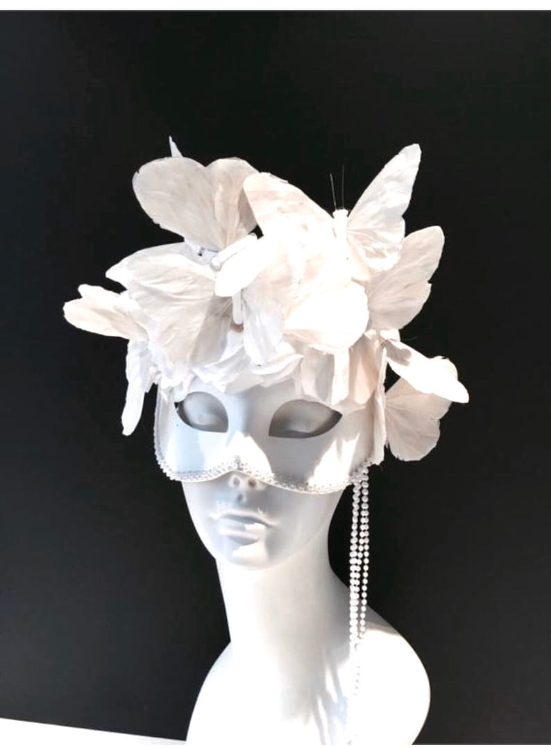 Puede incluir: Una m&aacute;scara blanca de carnaval con adornos de mariposas y flores. La m&aacute;scara presenta un ribete de cuentas y una hebra de perlas colgando del lateral. Se exhibe sobre una cabeza de maniqu&iacute; blanca sobre fondo negro.