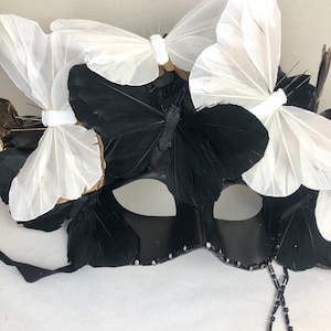 Puede incluir: Una m&aacute;scara negra de carnaval adornada con detalles de mariposas blancas y negras. La m&aacute;scara presenta una cuerda de cuentas y est&aacute; adornada con peque&ntilde;os pedrer&iacute;a brillantes a lo largo del borde inferior.