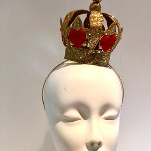 May include: A gold glitter crown headband featuring a series of red velvet heart-shaped appliques around the base. The crown has a central point topped with a small gold sphere and is attached to a thin, matching gold glitter-covered headband.