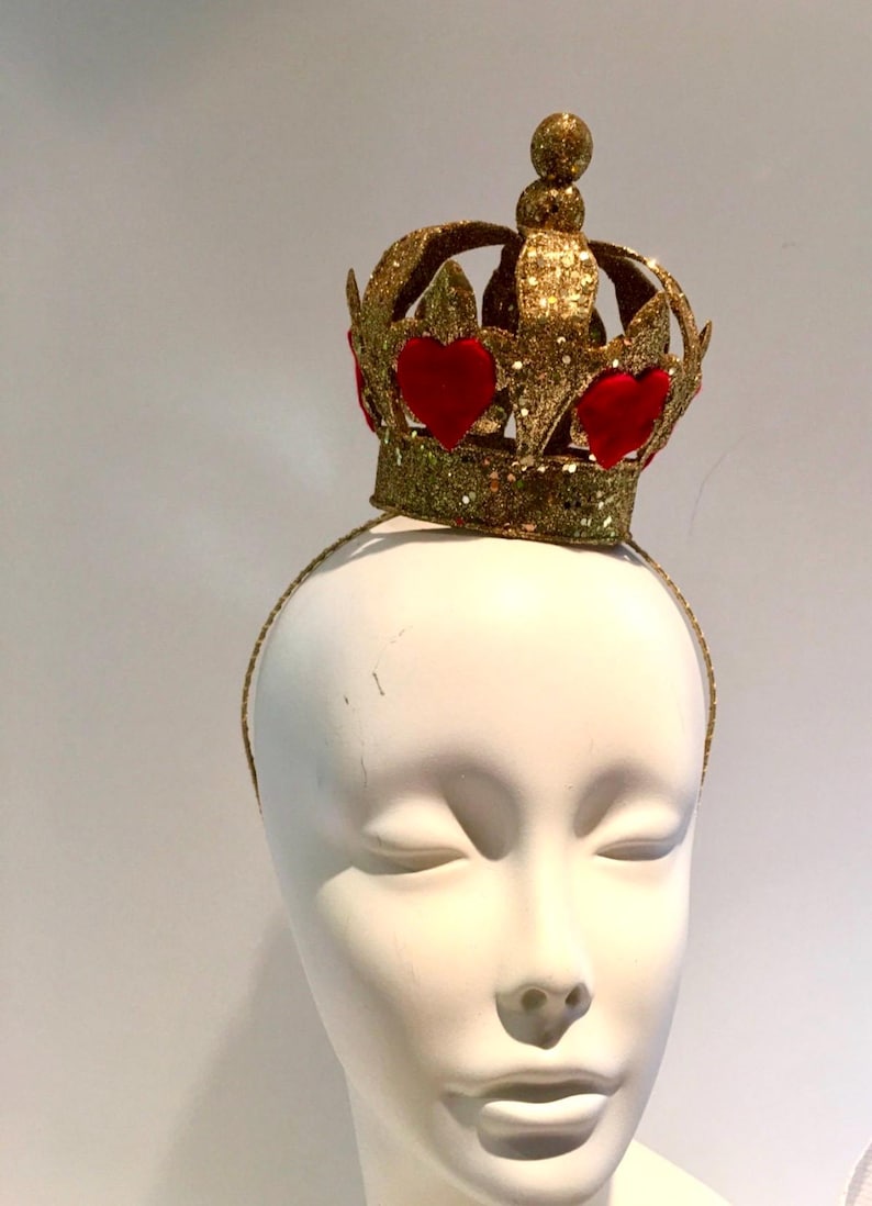 May include: A gold glitter crown headband featuring a series of red velvet heart-shaped appliques around the base. The crown has a central point topped with a small gold sphere and is attached to a thin, matching gold glitter-covered headband.